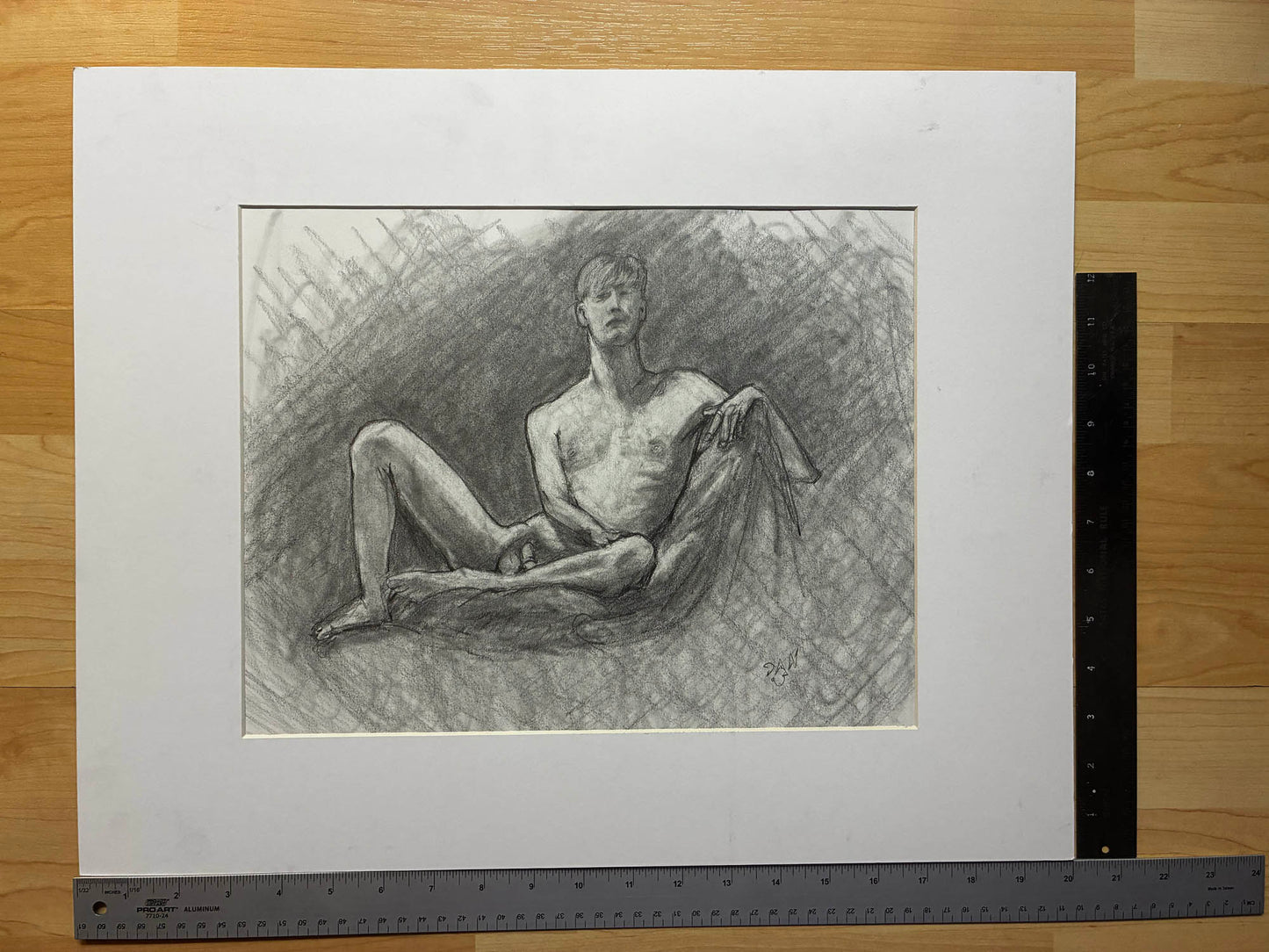 Pencil drawing of a nude figure on a white mat with a ruler for scale on a wooden surface.