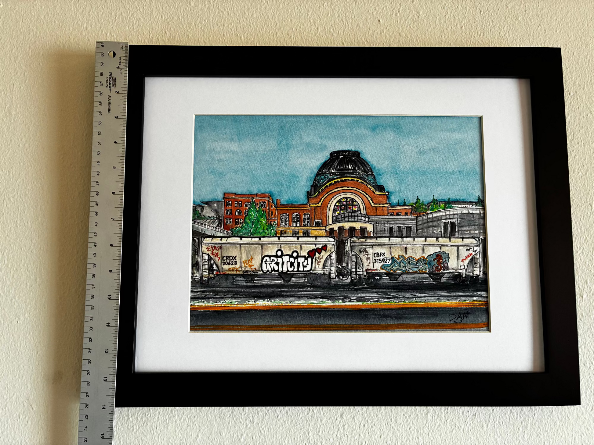 Framed artwork of a cityscape with a ruler for scale on a beige wall.