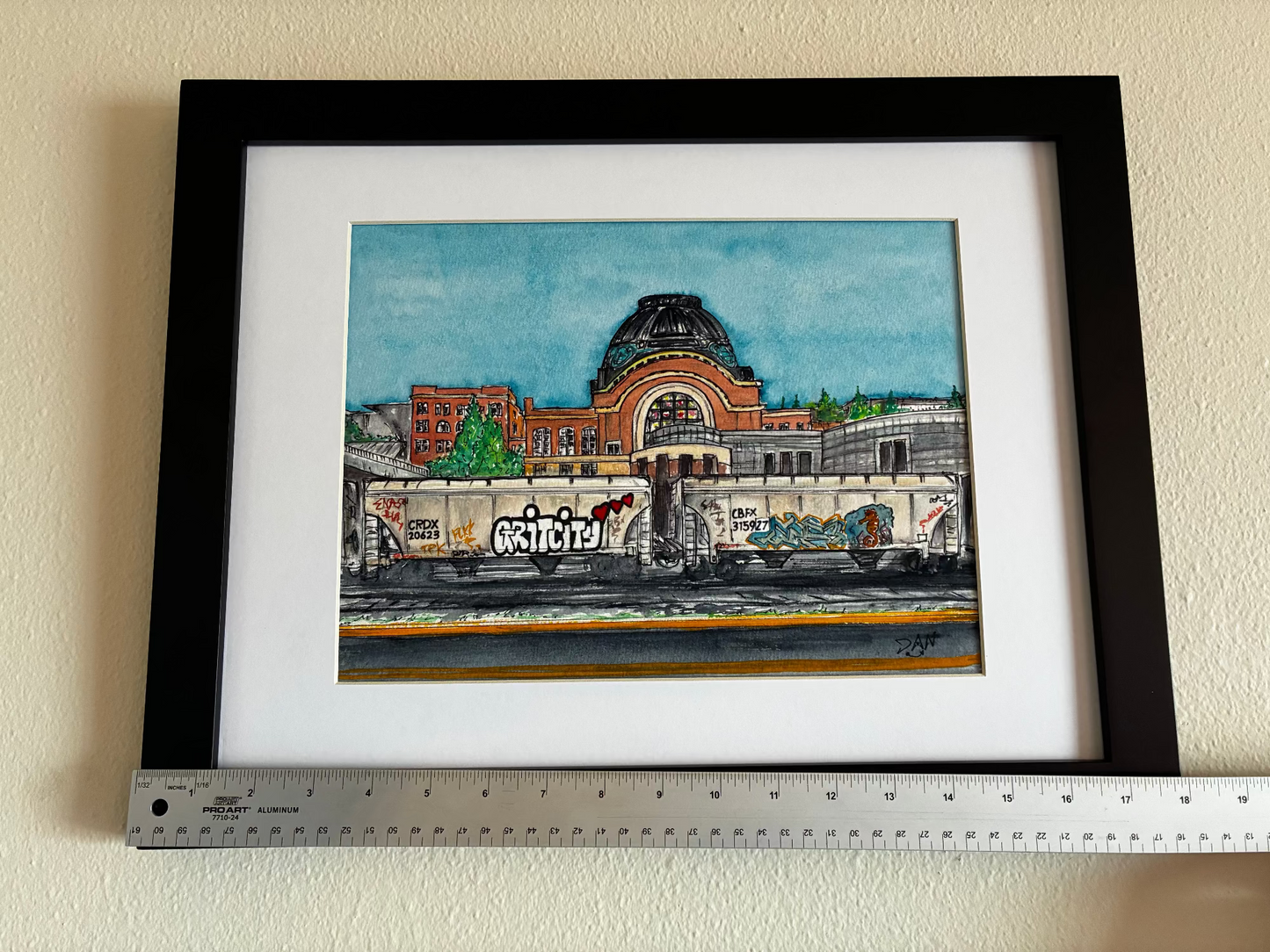 Framed artwork of a building with graffiti on a white wall, ruler at the bottom for scale.