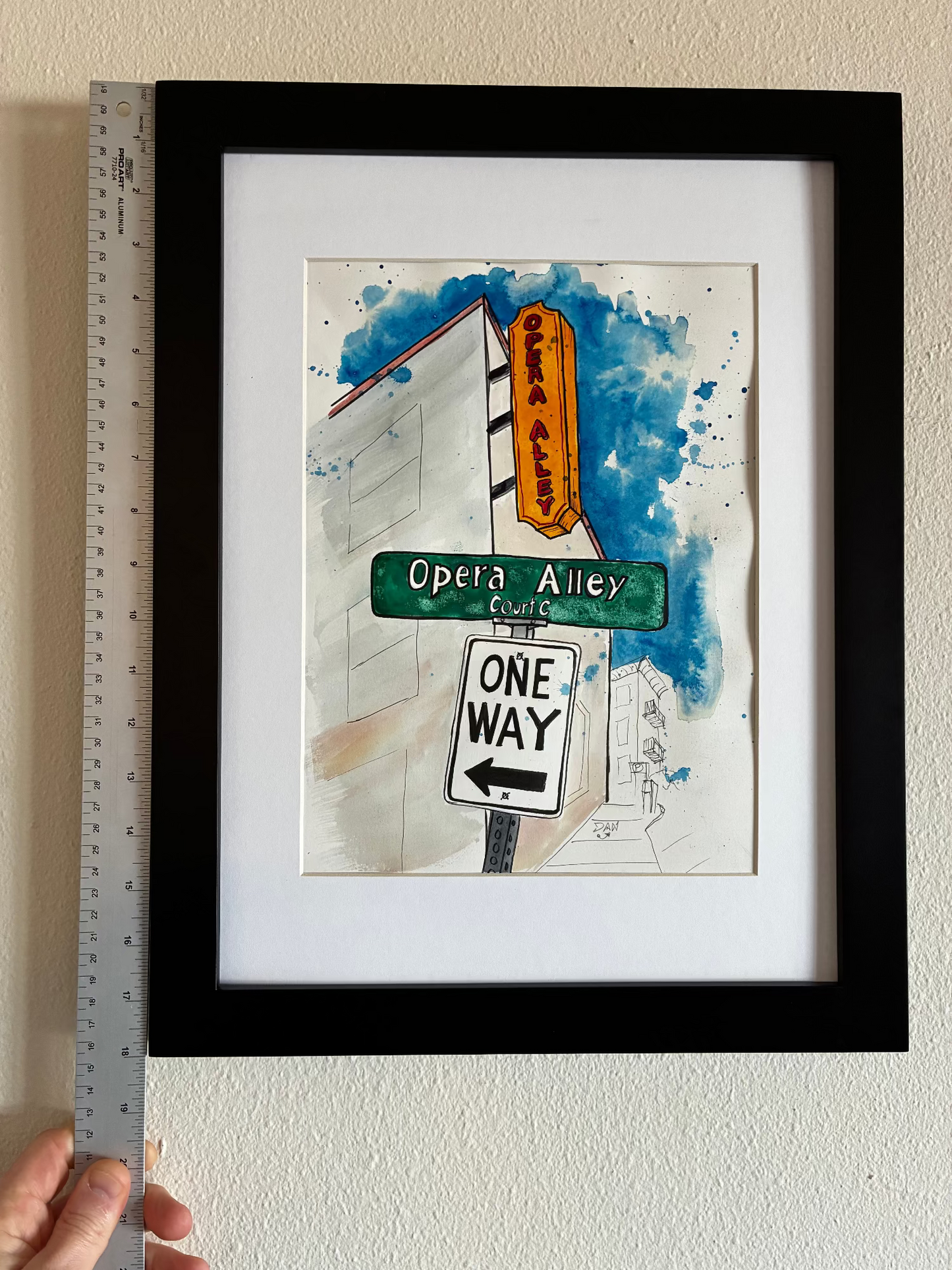 Framed artwork of street signs with a ruler for scale on a white wall.