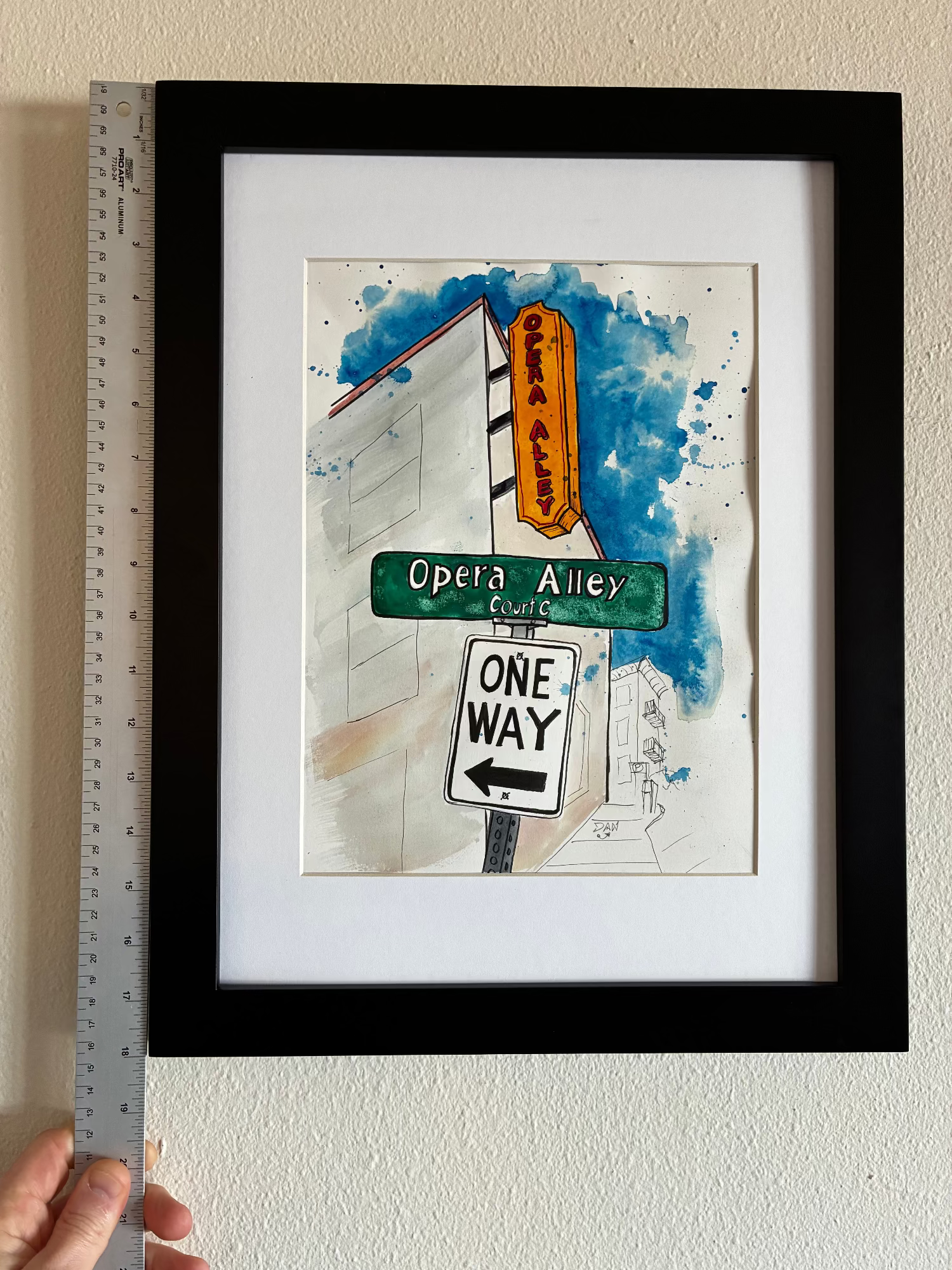 Framed artwork of street signs with a ruler for scale on a white wall.