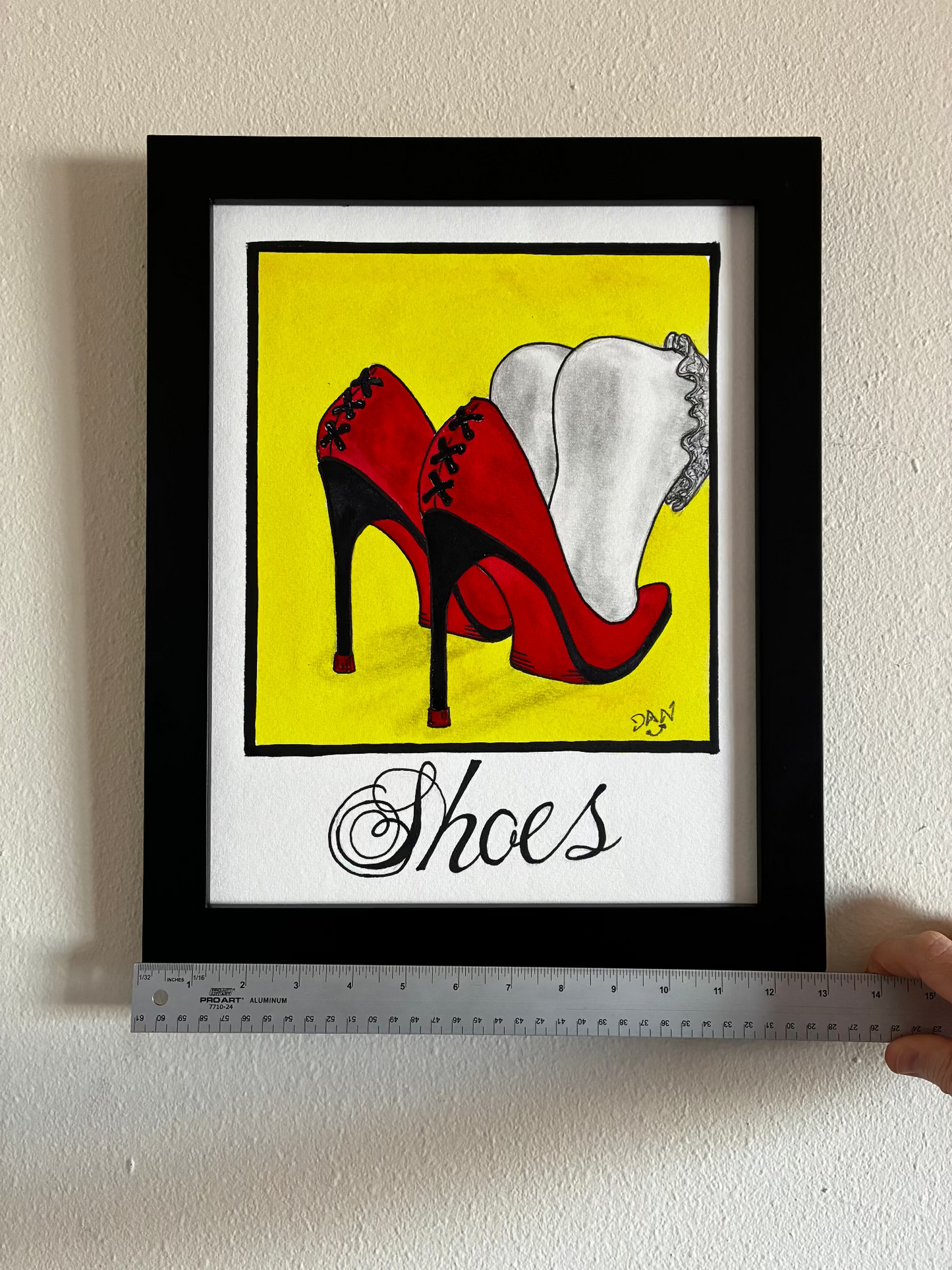 Framed artwork of red high heels on a yellow background with 'Shoes' text, held by a person with a ruler for scale.