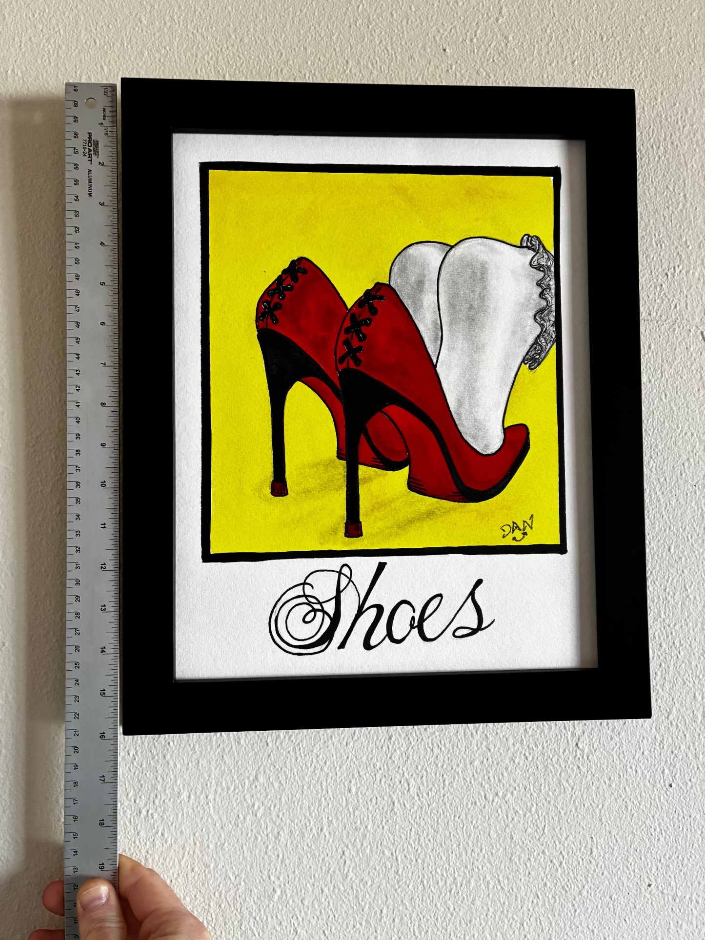 Framed artwork of red high-heeled shoes on a yellow background with 'Shoes' text, held up against a wall with a ruler for scale.