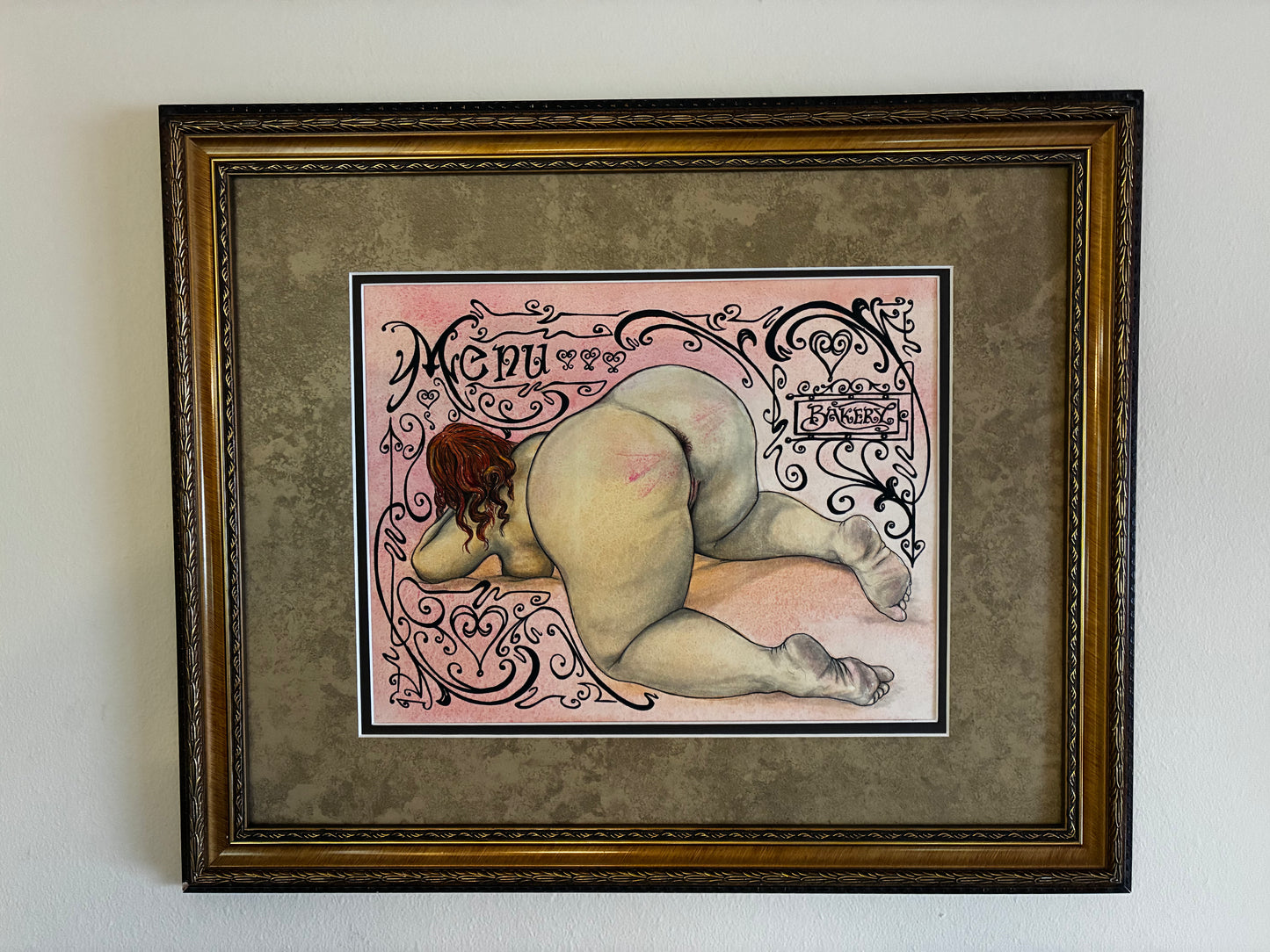 Framed artwork of a nude figure with decorative elements on a plain wall.