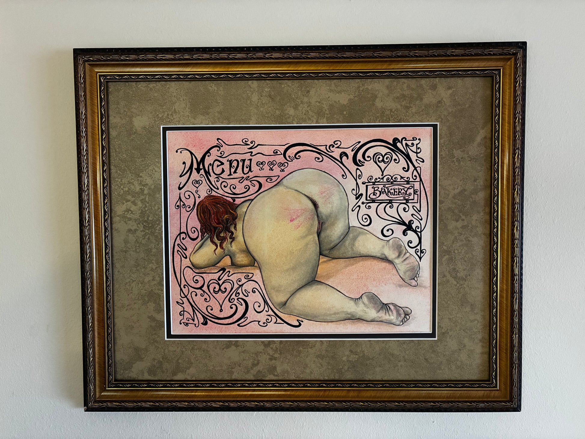 Framed artwork of a nude figure with decorative elements on a plain wall.